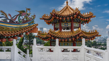 The beautiful tower of a Chinese Thean Hou Temple against a blue sky and clouds. Elegantly curved roof, bright lanterns, carved decorations. In the foreground there is a white stone railing. Malaysia.