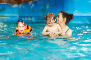 Joyful young mother with cute little sons has fun swimming and playing in large pool with clear...