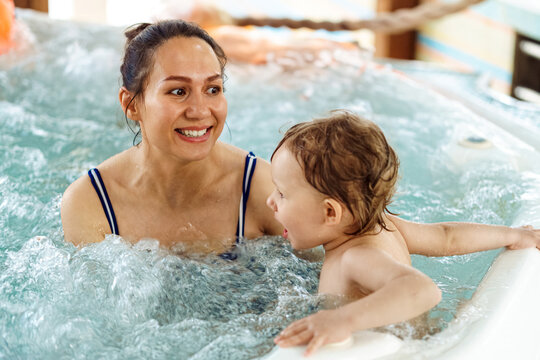 Cheerful woman with little child swim together in whirlpool bath. Happy mother and baby enjoy bathing in water with swirling bubbles at aquapark