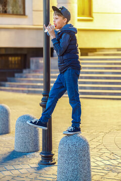 Playful boy eats ice cream standing on stone pillar in evening city. Kid licks sundae in cone having fun on street. Urban lifestyle and street food