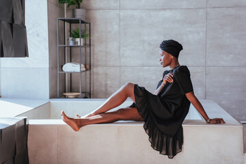 Lonely African girl in traditional clothes  and black turban sitting on bath at bathroom t home in sad mood, frustrated young Brazilian woman disappointed.