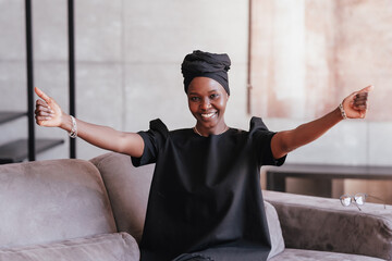 Cheerful African girl in black turban spreads hands looks at camera welcoming to hug sitting on...