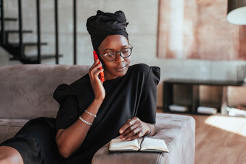 Focused African girl in black turban, glasses sitting on couch talks by phone  looks at opened diary remote working home. Young Brazilian entrepreneur makes online order. - obrazy, fototapety, plakaty
