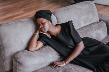 Exhausted African girl in black turban and black dress laying on couch eyes closed having nap,...