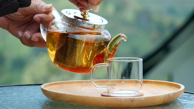A hand pouring tea from glass teapot on wooden serving tray, hands pouring tea from teapot, Cropped shot of pouring tea in traditional chinese tea ware