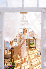 Fototapeta na wymiar Vertical portrait of charming blonde female in beautiful dress standing inside of summer gazebo house on sunny day, smiling looking at camera with happy expression. Concept of summertime vacation.