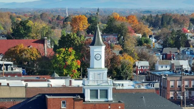 A white clock tower stands tall amid autumn-colored trees in a serene town. Aerial zoom lens.