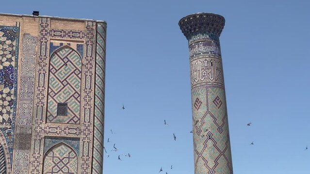 Birds circle Ulugh Bek Madrassah's minaret in Registan Square, Samarkand, Uzbekistan. Marvel at Intricate Mosaics, Persian-Timurid Fusion, and Masterful Craftsmanship in this UNESCO Heritage Site