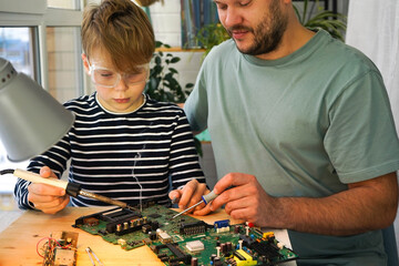 Father with his son spend time together. Close-up of a man teaching his boy at home to solder computer spare parts. Education moment during parenthood. Togetherness concept.