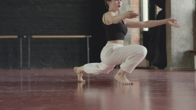 Young Caucasian barefoot woman wearing white relaxed joggers and black crop top showing contemporary choreographic improvisation, dancing on parquet floor in modern dance studio