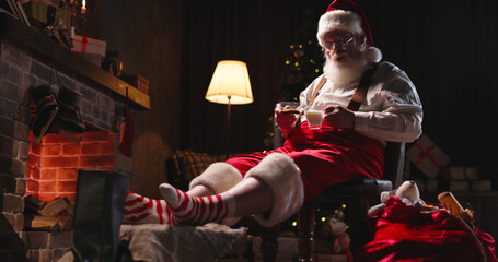 Tired Santa Claus sleeping near fireplace, holding cookies and cup with milk. Overworked Mr Claus...
