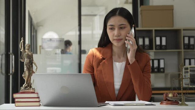 Young attractive Asian female lawyer in formal suit works on tablet with laptop, legal books, and justice scale on her desk.
