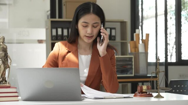 Young attractive Asian female lawyer in formal suit works on tablet with laptop, legal books, and justice scale on her desk.