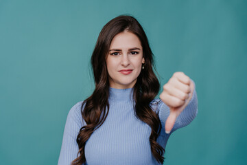 Brunette caucasian girl with wavy hair looks at camera shows thumb down gesture, negative decision standing against turquoise background. Don’t do this, its not good choice. - obrazy, fototapety, plakaty