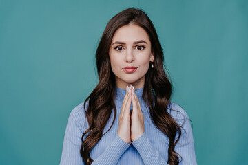 Brunette girl with wavy hair holds hands in prayer gesture looks at camera with hopeful face...