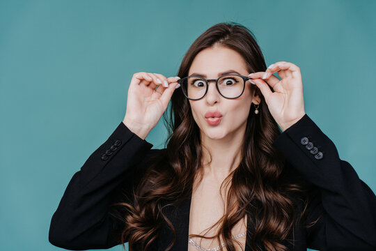 Curious brunette girl in puts on glasses looks at camera in surprise with wide opened eyes wears black suit standing against turquoise studio background. Mockup, sale enormous discount.