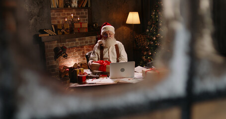 Bearded Santa Claus using laptop outside view through window, talking on video call, holding New...