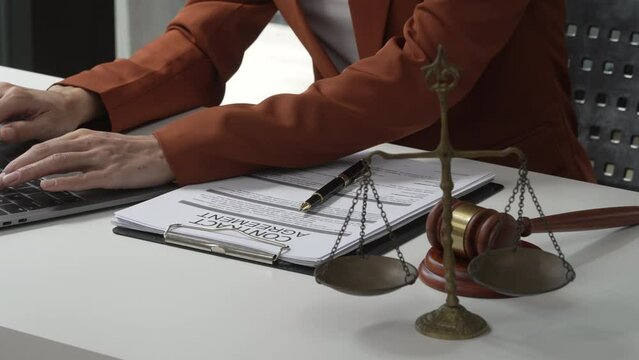 Young attractive Asian female lawyer in formal suit works on tablet with laptop, legal books, and justice scale on her desk.