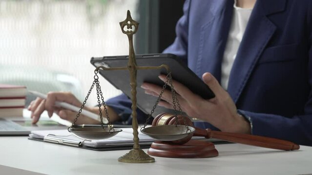 Young attractive Asian female lawyer in formal suit works on tablet with laptop, legal books, and justice scale on her desk.