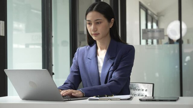 Young attractive Asian female lawyer in formal suit works on tablet with laptop, legal books, and justice scale on her desk.