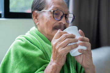 Selective of one Asian male senior man wearing glasses and sit on sofa bed at home hold cup of tea...