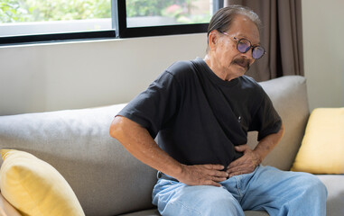 Senior Asian man sitting on sofa having suffering from stomach ache holding his stomach pain,...