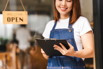 Happy excited attractive young Asian woman in denim apron, received online order in coffee shop with an 'Open' sign.