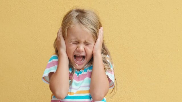 Unhappy sad angry little child girl close eyes and cover ears by hands being annoyed with loud noise, isolated yellow studio background. Children emotions. Child dislike loud sounds