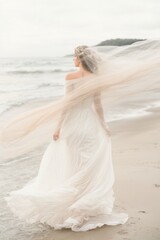 Fototapeta na wymiar A happy, rejoicing young woman bride in a long white wedding dress stands on the seashore, a sandy beach. The veil curls and flies in the wind, waves and a white sky on the background.