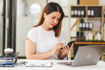 Fototapeta premium Cheerful Asian woman confidently uses mobile phone, tablet with laptop and notebooks on the desk in a bright office.