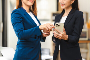 Two middle aged and young Asian female executives in formal suits review bar chart, discussing business strategies in office setting, senior executives or directors in advertising or public relations
