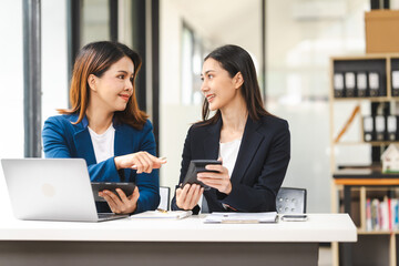 Two middle aged and young Asian female executives in formal suits review bar chart, discussing business strategies in office setting, senior executives or directors in advertising or public relations