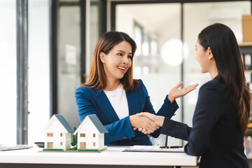 Two Asian real estate agents in formal suits discuss a contract with house models on the table,...
