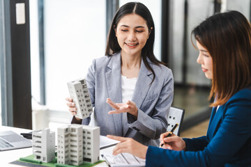 Two middle aged and young Asian business woman in formal suits discuss over model buildings, on...