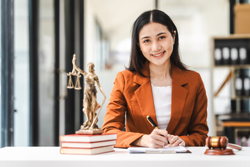 Young attractive Asian female lawyer in formal suit works on tablet with laptop, legal books, and...