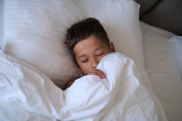 A young boy sleeps peacefully, enveloped in soft white bedding, with gentle morning light caressing the scene. This authentic image resonates with the universal need for restful sleep and the growing