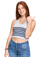 Young read head woman wearing casual clothes smiling with happy face looking and pointing to the side with thumb up.