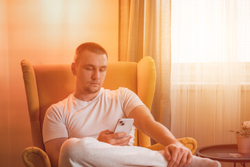 Young blonde man at home on the sofa with mobile phone.  He relaxes on a couch in his comfortable home, using his cellphone to check social media and chat with his friends.