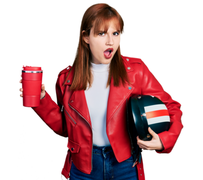 Redhead young woman holding motorcycle helmet and take away coffee in shock face, looking skeptical and sarcastic, surprised with open mouth