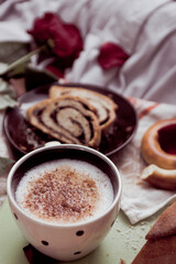 Breakfast in bed close-up, cappuccino in a light cup and pastries with poppy seeds on a board, against the background of a kitchen towel, a red rose and blanket