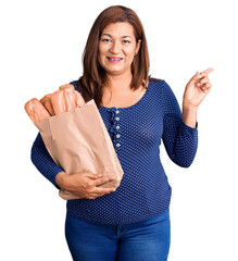Middle age latin woman holding paper bag with bread smiling happy pointing with hand and finger to...