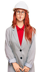 Young redhead woman wearing architect hardhat making fish face with lips, crazy and comical gesture. funny expression.