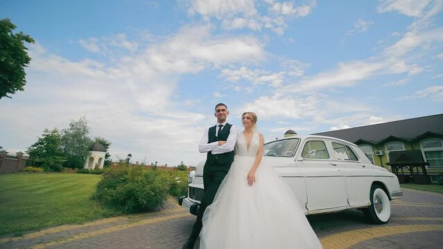 The newlyweds stand near a retro car and kiss against the background of the blue sky. The wedding day of the newlyweds in love, a camera runs over them, they pose for the camera.
