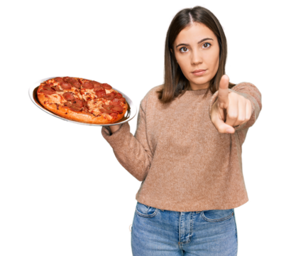 Young beautiful woman holding italian pizza pointing with finger to the camera and to you, confident gesture looking serious