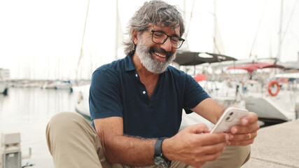 Happy senior man in glasses sits in the port on yacht background uses smartphone, looks at funny...