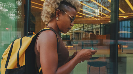 Smiling young woman in glasses, wearing brown top with backpack on her shoulders, walking down the street with smartphone, texting, chatting with friends on social media