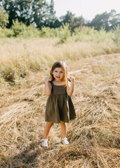 Little girl standing outdoors on a field on summer day, wearing a linen dress, looking at camera.