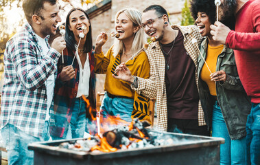 Happy friends roasting marshmallows over campfire - Group of young people enjoying garden party at...
