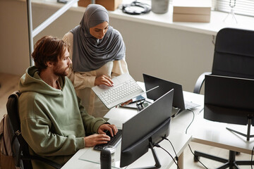 High angle portrait of female computer programmer wearing headscarf in office and showing laptop screen to colleague during code review