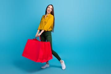 Full length photo of sweet dreamy woman dressed yellow shirt holding shoppers looking empty space isolated blue color background